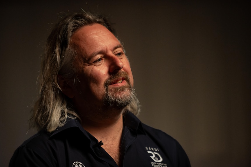 A bearded man with long, fair hair wears a dark shirt in a dimly-lit room.