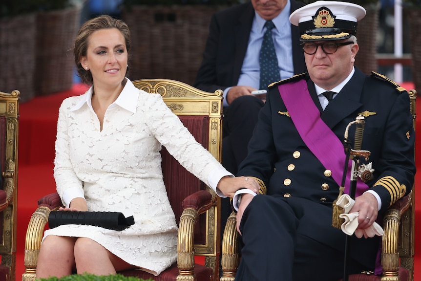 A couple sit in golden chairs, holding hands. The man is wearing a military uniform.