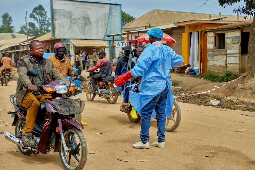 A man in blue protective gear stands in the middle of the street, blocking people on motorcycles.