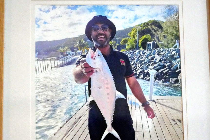 A framed photograph of a smiling man in a hat holding up a fish while standing on a pier.
