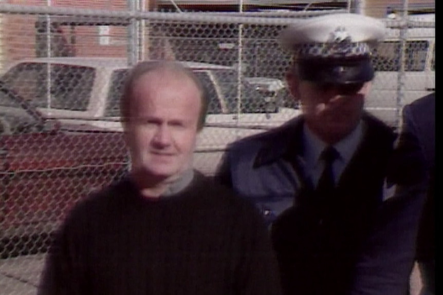 A balding man walks next to a uniformed police officer.