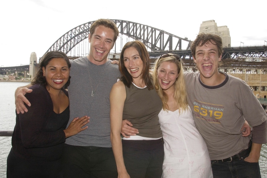 Three women and two men stand laughing and smiling with the Sydney Harbour Bridge in background.