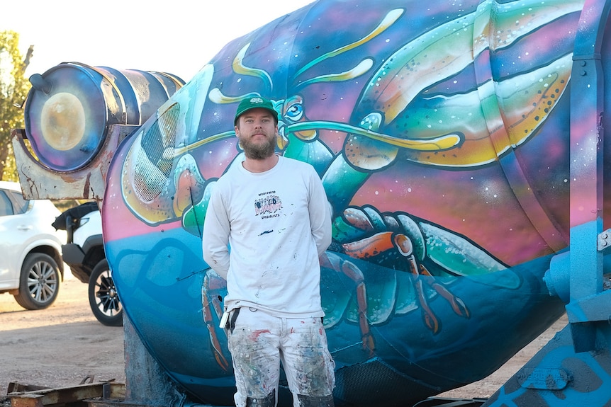 A man in a paint-flecked white clothes stands in front of an artwork