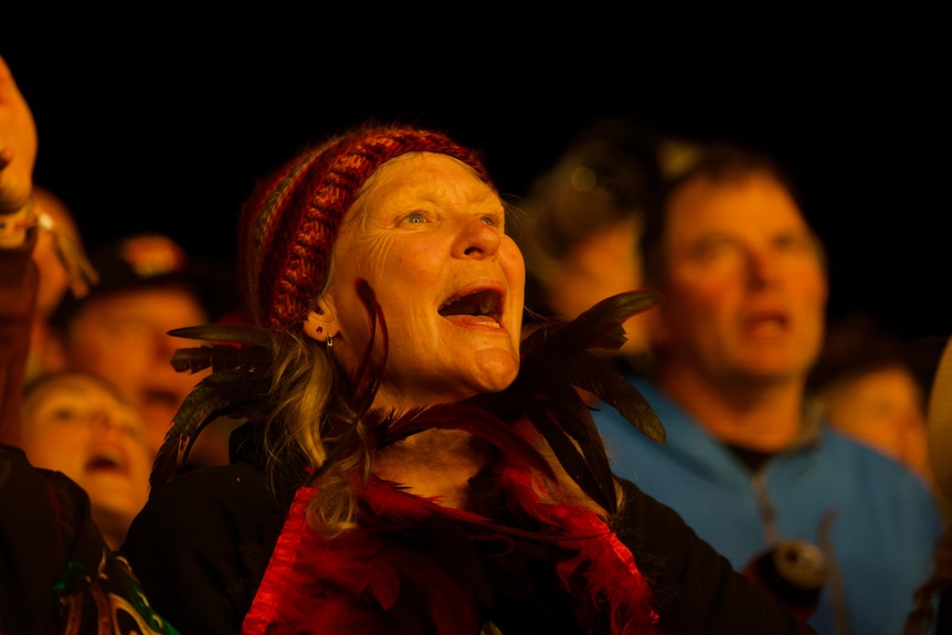 A woman wearing a beanie and a feathered collar sings in a crowd