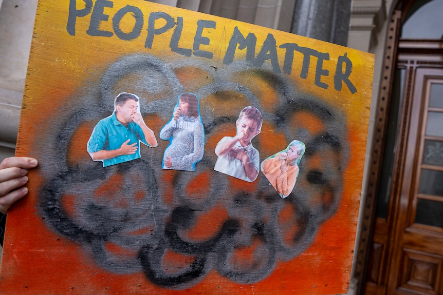 A person holds a placard showing coughing children outside the doors of parliament reading 'people matter'