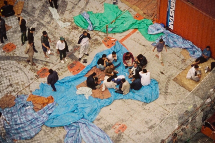 An aerial shot of people sitting on tarpaulins in the concrete deck of a ship.