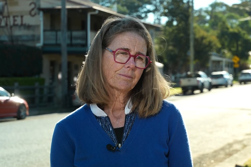 A woman with shoulder-length hair and glasses stands in front of a town street.