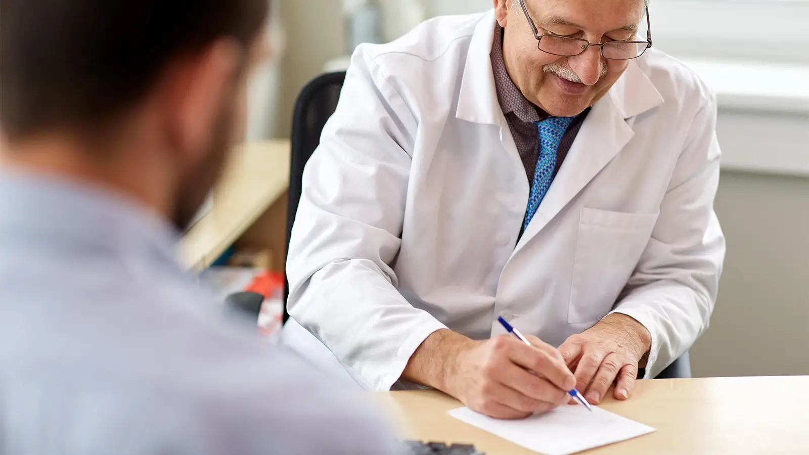 A photo of a senior male physician writing a prescription for his male patient.