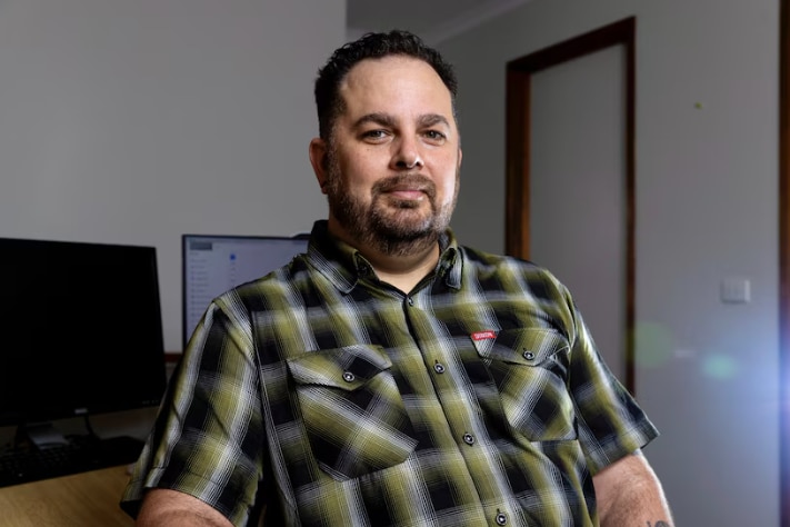 A man with a green checked shirt sits in front of his desk.