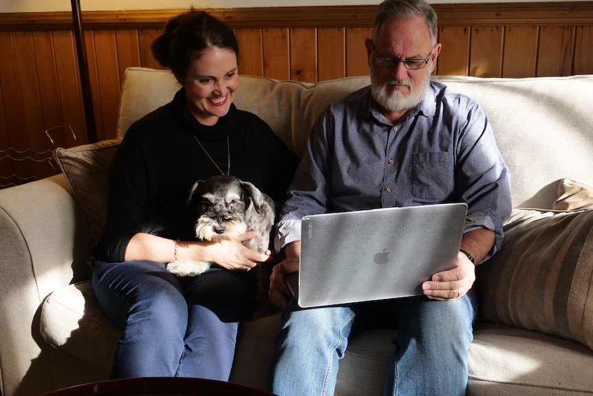 A man and woman, the latter with a cat on her lap, sit on a couch looking at a laptop.