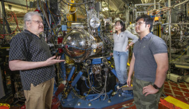 Three researchers meeting in a lab with a complex setup of large metallic instruments.