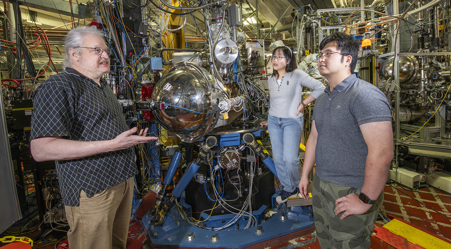 Three researchers meeting in a lab with a complex setup of large metallic instruments.