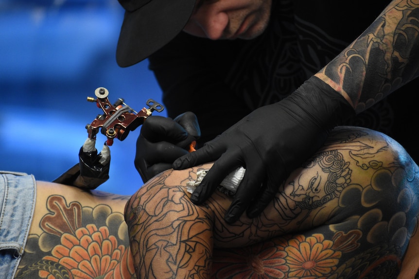 A man in a black cap leans over a person's midriff and applies an intricate tattoo to their skin