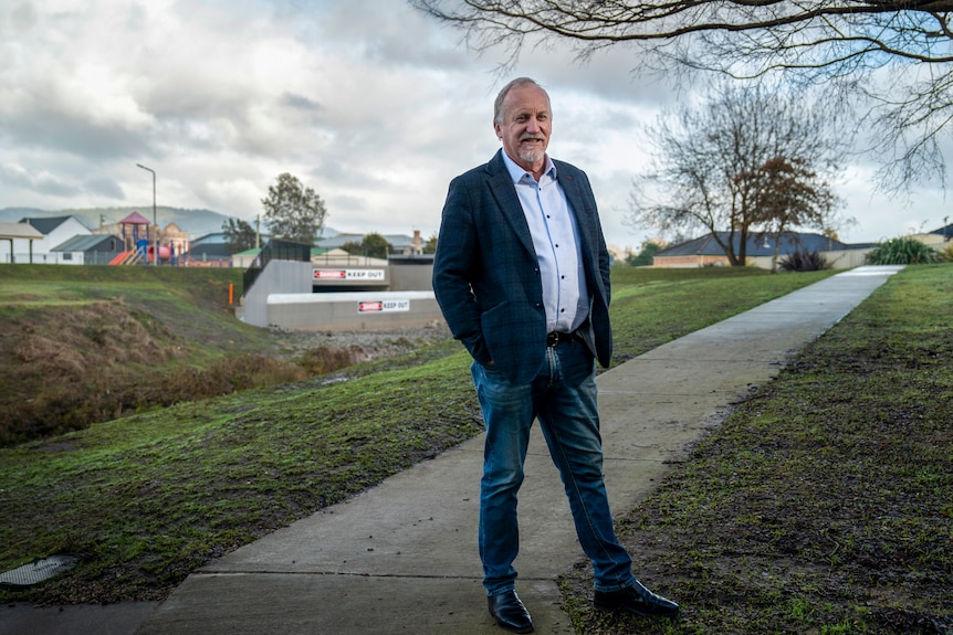 An older gentleman in a navy suit and jeans stands in a park with a path, creek and concrete structures behind him.