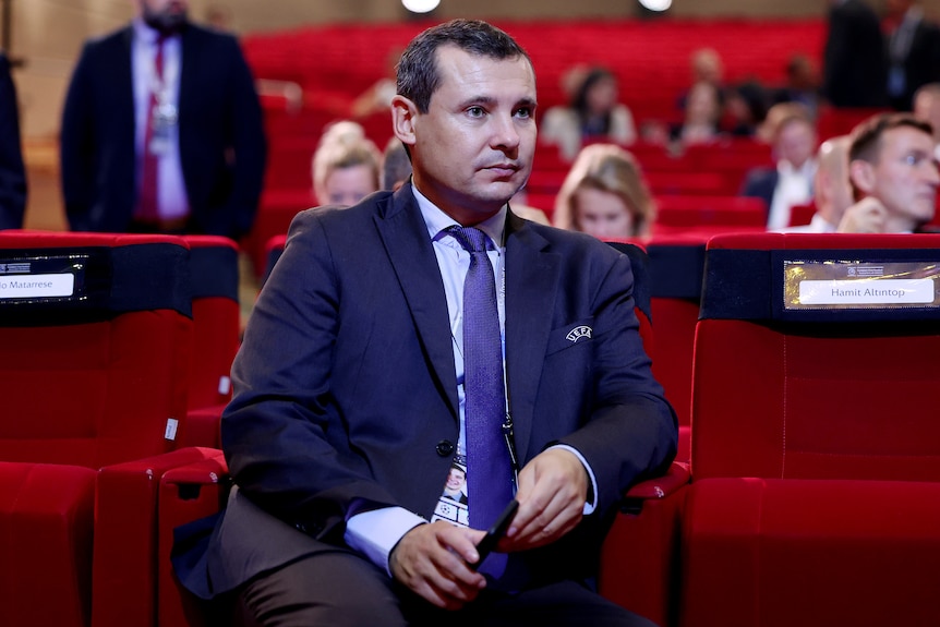 A man in a suit sits in a theatre chair.
