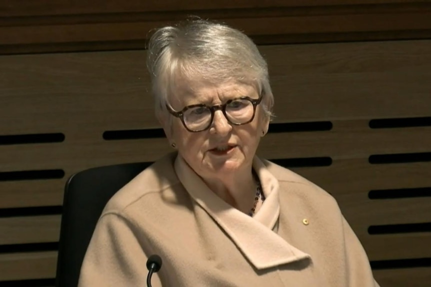 Woman in caramel jacket and wood panelling behind her