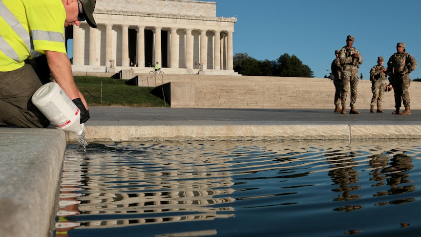 A man wearing a hi-vis shirt kneels by the Reflecting Pool and empties a bottle of bleach into the water