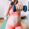 A photograph of an unrecognizable pregnant woman exercising with dumbbells in her living room at home.