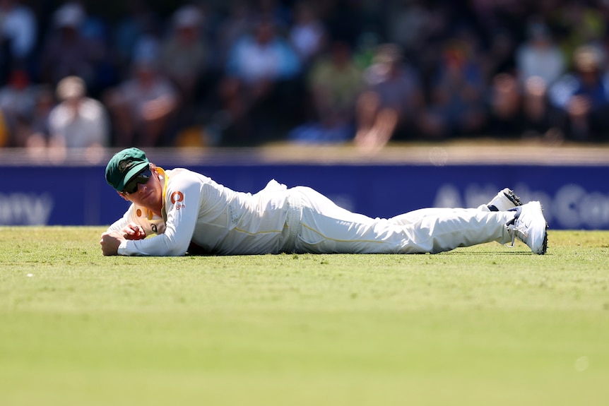 A cricketer in white lays down on the grass while looking concerned