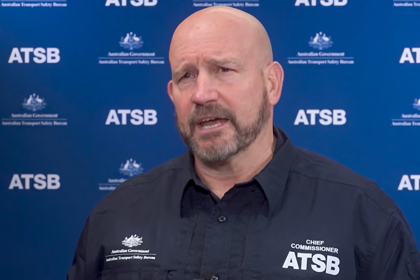 Man with bald head, dark facial hair, dark shirt, behind ATSB signage