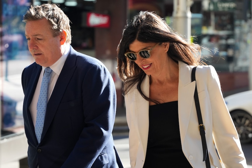 Smiling woman with dark hair wearing sunglasses and white jacket with black shirt in the street