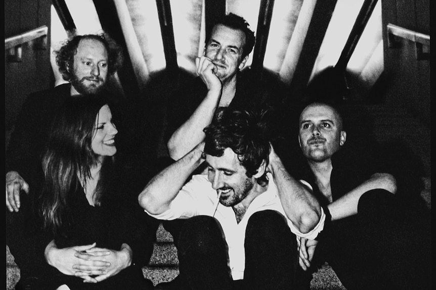 Black and white press shot of The Drones' six members sitting on venue steps