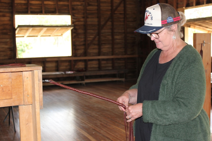 A woman in a green cardigan, glasses and cap pulls on leather strands and plaits them.