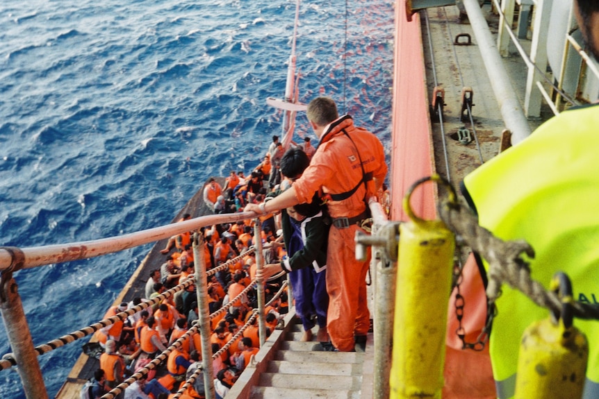 People in red life jackets climb from a small wooden boat up some stairs to the side of a freighter.