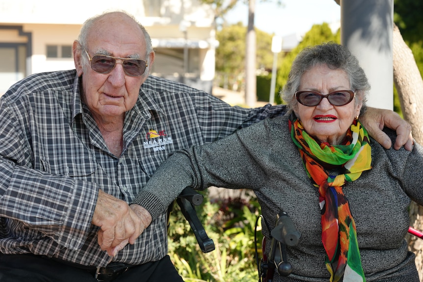 An older couple sit in walking frames hand in hand.
