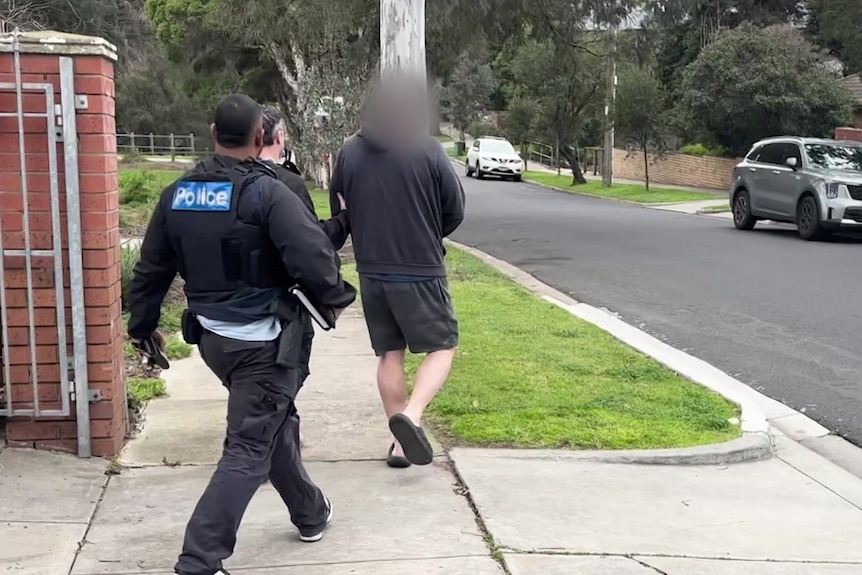 A man wearing a black jumper, shorts and thongs is led away by two police officers.