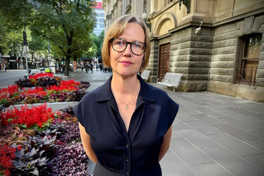 A woman with short hair and glasses stands in a street.