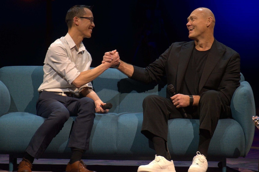 Eddie Woo and Michael Klim are seated on a couch on a stage, grasping hands and smiling at each other.