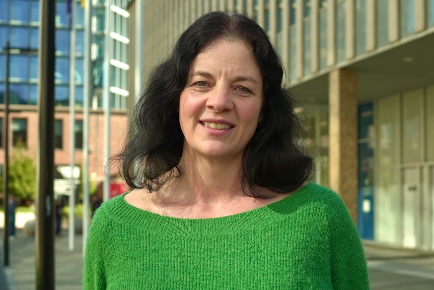 A woman with long dark hair in a bright green jumper stands smiling outside a building with gold-tiled columns.