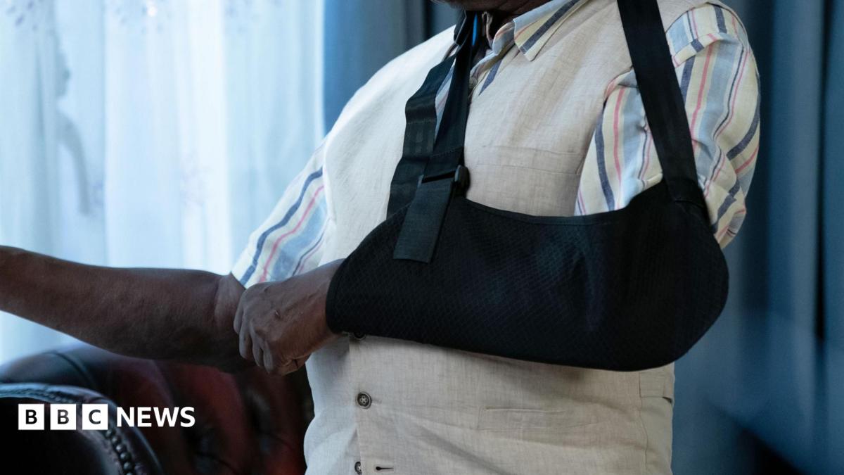 A close-up image of  man's left arm in a black sling. He is also wearing a white waistcoat and striped, short-sleeved shirt.