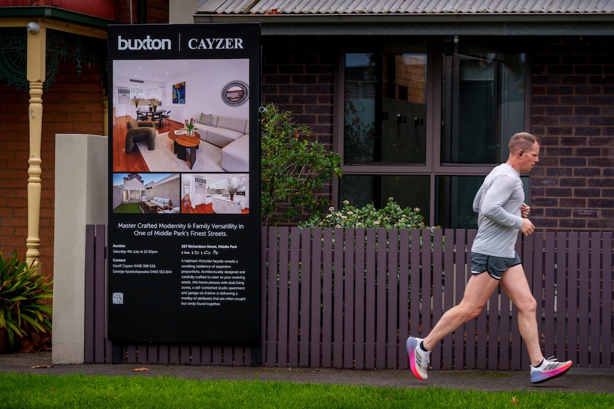 A man jogs past a brown brick house that has a large sign attached to the fence indicating it is for sale.