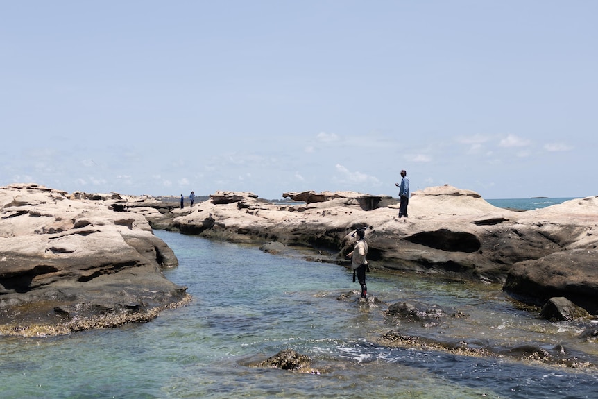 A team of people standing on rocky shore, some are atop the rock and one has his feet in the ocean.
