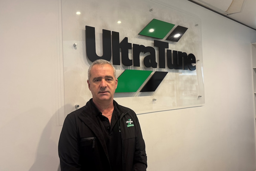 A grey-haired man in a dark shirt stands in front of the Ultra Tune logo in an office foyer.