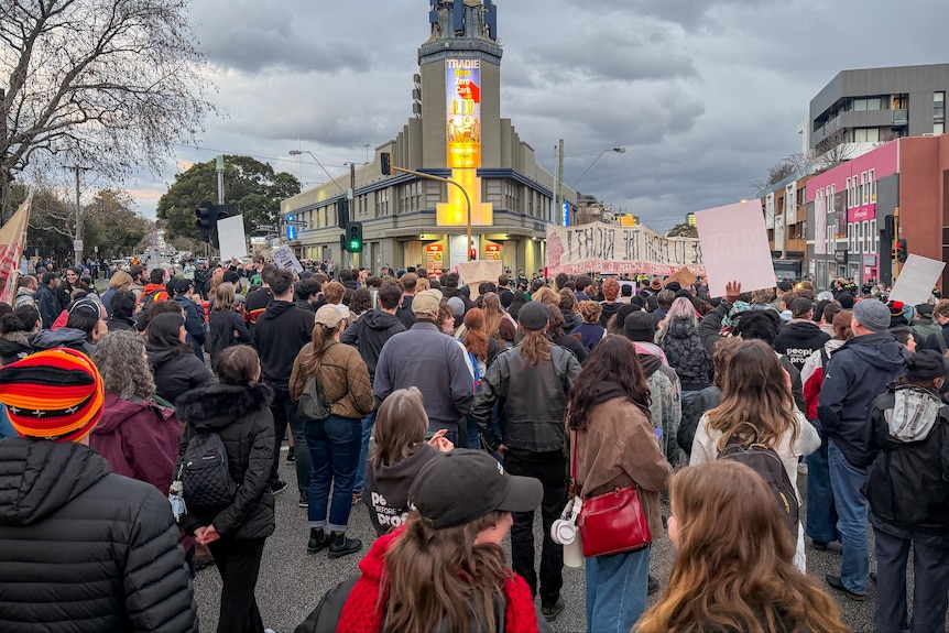 A large crowd of people, some with banners, cover the road outside a hotel at dusk.