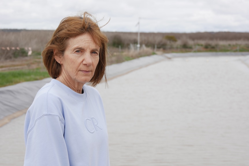 A woman with brown hair and a blue jumper stands in front of a river looking solemn
