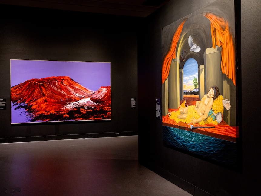Two paintings hang in an exhibition space: one of a red and purple landscape, the other of an Indigenous man reclining.