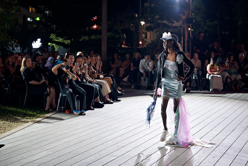 A model in a turquoise outfit walking down a runway, at a night-time fashion show.