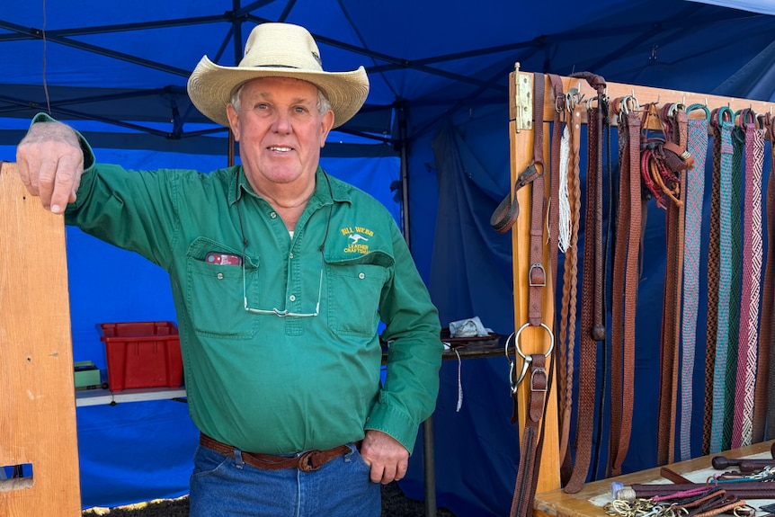 A man wearing a green shirt and a white cowboy hat smiles at the camera while leaning with one arm on a timber stand..