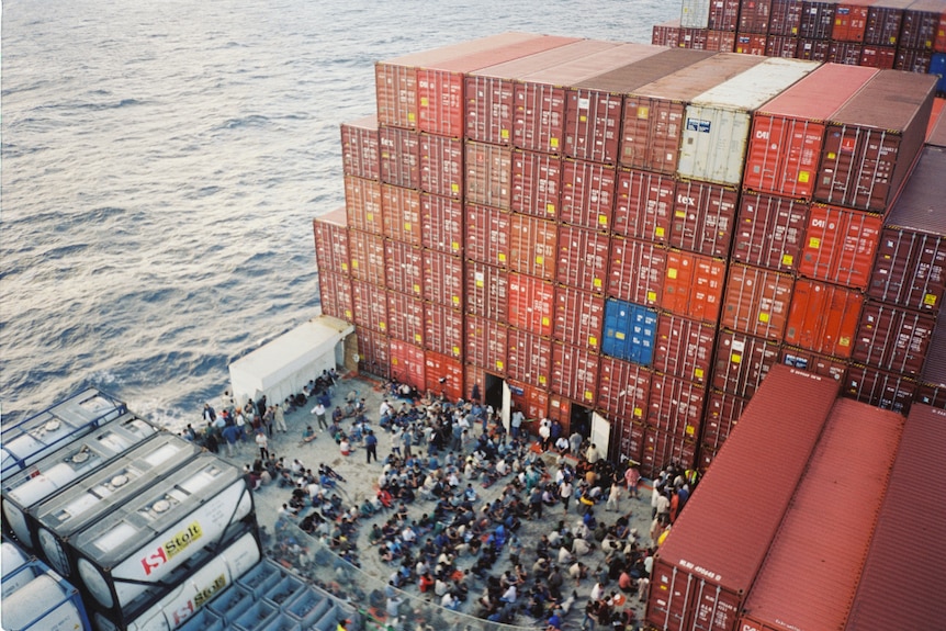 An overhead shot of a crowd of people sitting and standing on the deck of a large freight ship.