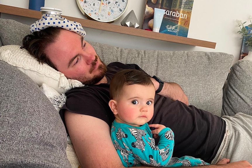 A man lying on a couch with an ice bag on his head and a baby next to him.