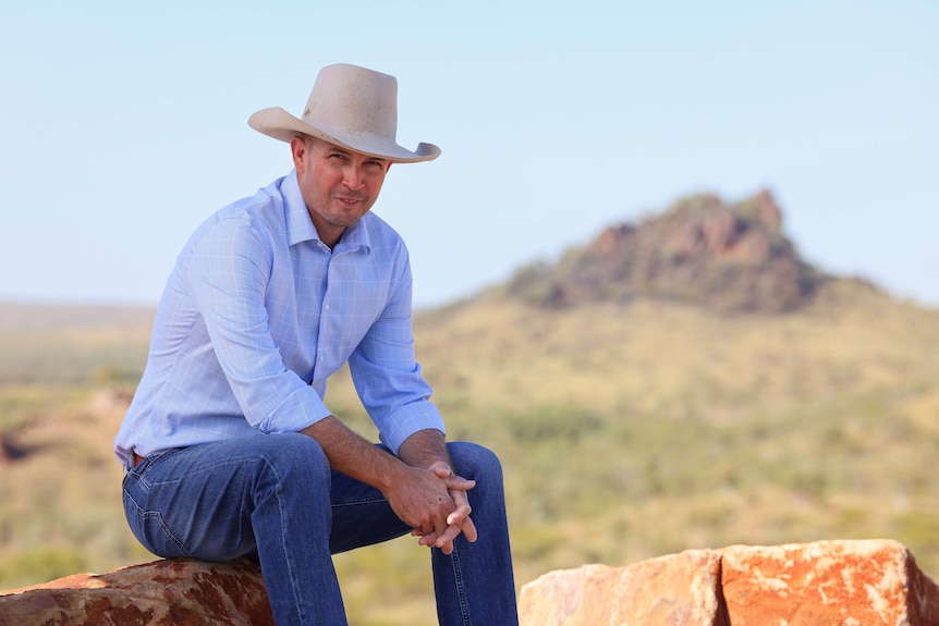 A man in outback business attire sits on a rock with a rocky landscape behind him.