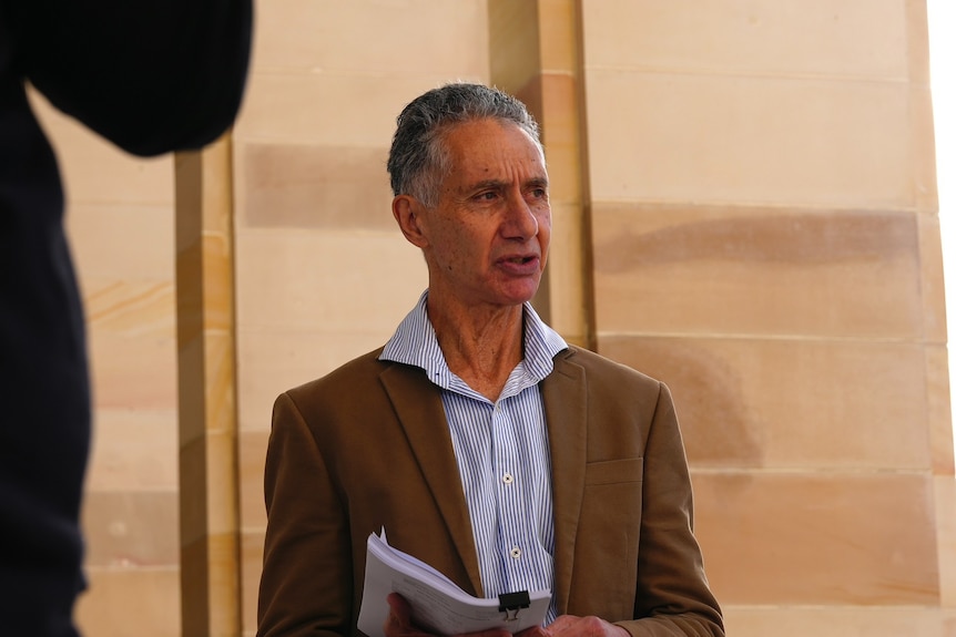 A mid-shot of a man wearing a brown suit jacket and light-coloured shirt