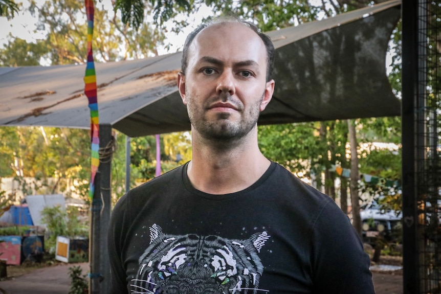 A man with a serious facial expression looks at the camera, he has facial hair and is wearing a black t-shirt.