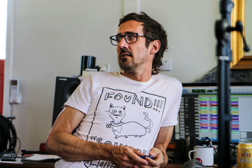 Gareth Liddiard wearing black-framed seeing glasses looks across room, sitting in front of production computer.