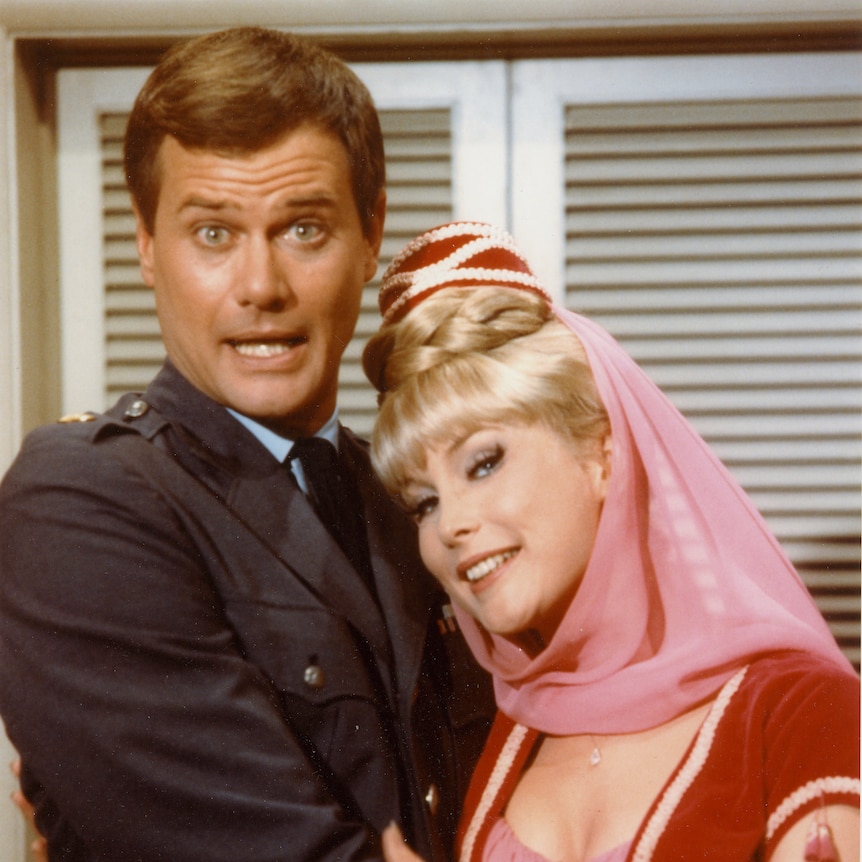 A male actor in a suit with a female actor dressed as a genie with long blonde flowing hair and pink headwear