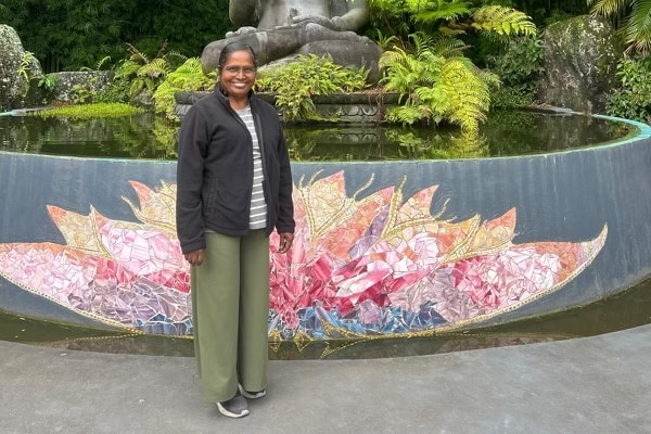Kanagamah Sundrasan in a black puffer jacket, green pants, smiling in front of a spiritual monument with a lotus.